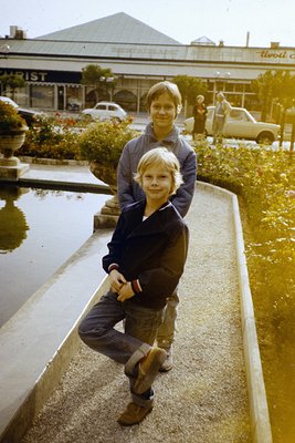 Two boys pose on a concrete ledge beside a small water feature in a mid-20th-century urban setting. The older boy (front) wea...