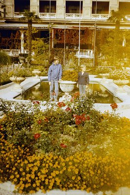 Two men pose in a sunlit courtyard garden with a central fountain and blooming roses, framed by classical architecture. Warm ...