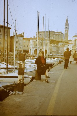Vintage sepia-toned waterfront scene featuring a woman in a red coat and white stockings seated on a dock post, likely **Pula...