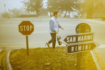 Vintage sepia-toned street scene at a road junction near San Marino/Rimini border. A man in a light jacket and dark pants wal...