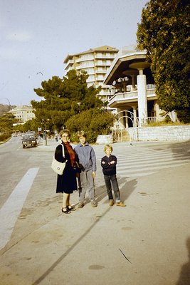 Three individuals pose on a wide, curved seaside promenade beside a mid-century resort-style building with arched balconies a...