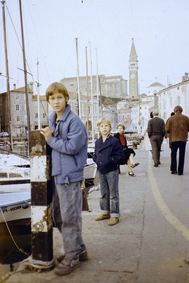 1970s coastal promenade with two boys in casual attire (blue jacket, jeans) near a docked boat. Historic European architectur...