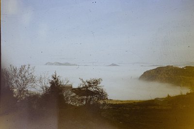 Vintage sepia-toned coastal scene with misty horizon and distant islands. A lone building sits on rocky terrain near water’s ...