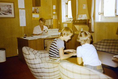 Vintage office scene featuring a seated female administrator behind a wooden desk with typewriters, surrounded by framed docu...