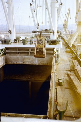 Mid-20th century industrial ship deck with exposed cargo hold. Rust-stained metal railings, pipes, and winches frame a large ...