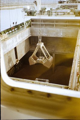 Industrial cargo hold mid-loading via overhead crane with clamshell bucket. Concrete bulkhead and metal decking visible. Like...