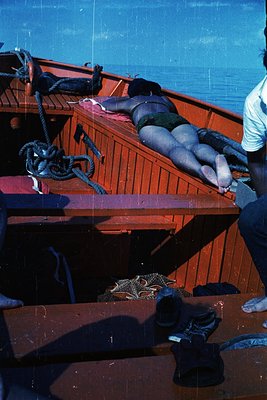 Vintage wooden fishing boat deck with two people sunbathing on a red-painted bench. Rope coils and worn shoes on deck suggest...