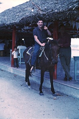 Man in 1970s-style denim jacket and jeans poses atop a saddled horse in a seaside resort setting. Thatched-roof pavilion and ...