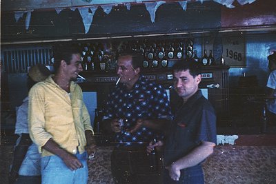 Vintage bar scene with three men in 1960s-70s attire. Centered: man in polka-dot shirt smoking, holding a drink. Left: man in...