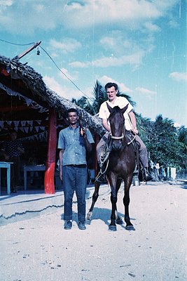 Vintage black-and-white photo of two men in rural setting: one standing beside a horse-drawn cart, the other mounted on a hor...