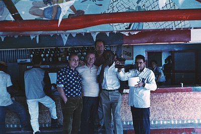 Vintage bar scene with four men posing behind a polished wooden counter, 1960s-70s. Decor includes a mural of a sailboat and ...