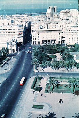 Aerial view of a mid-20th century Mediterranean cityscape, featuring Brutalist architecture and palm-lined boulevards. Promin...