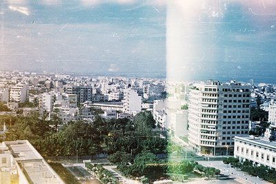 Vintage urban skyline shot through a yellowed window pane, revealing mid-rise concrete buildings and greenery. Likely 1960s–1...