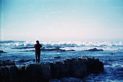 Vintage coastal scene featuring a lone figure in a red jacket standing on jagged rocks by crashing waves. Vibrant blue ocean ...