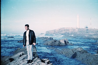 Man in 1970s-style suit poses on rocky coastline near lighthouse. Vibrant blue seascape contrasts with muted tones of his att...