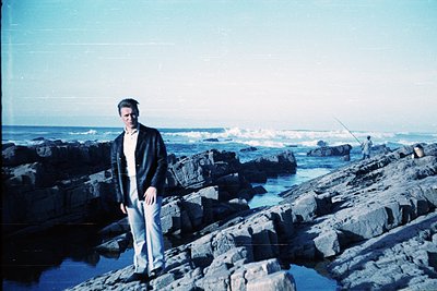 Man in formal attire (dark blazer, light trousers) stands on rugged coastal rocks by the sea, mid-20th century. Vintage polar...