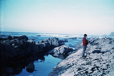 Vintage coastal fishing scene: lone angler in red jacket and light pants stands on rugged rocky shore, casting line toward oc...