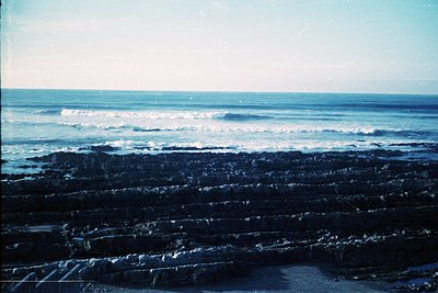 Vintage seaside shot of rugged, layered rock formations meeting the ocean. Waves crash against jagged cliffs under a clear sk...