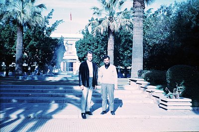 Two men pose on a wide, tiled plaza flanked by palm trees and classical architecture, likely a public square. The vintage sep...