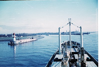 Vintage black-and-white ship deck view showing industrial port infrastructure. Foreground features a cargo vessel’s railings ...