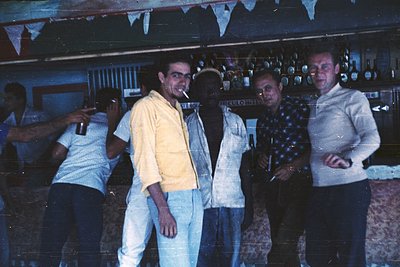 Vintage bar scene featuring four men in 1960s-70s attire—yellow button-up, light blue shirt, patterned sweater, and white jac...
