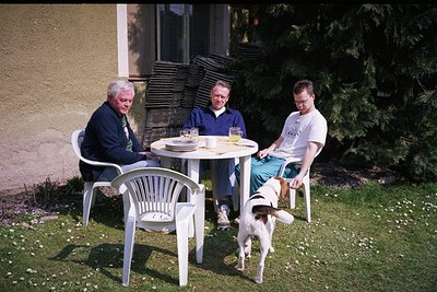 Three men seated at a small outdoor table in a garden, mid-1990s. Left: older man in dark blazer, white shirt, and tie. Cente...