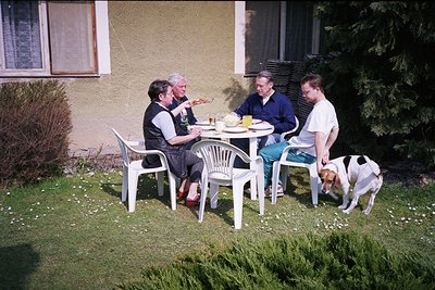 Four adults and a dog share a casual outdoor meal on white plastic chairs around a small round table in a residential backyar...