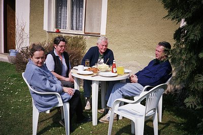 Four adults seated at a plastic table in a residential backyard, mid-1990s. Light-colored stucco house with white-framed wind...