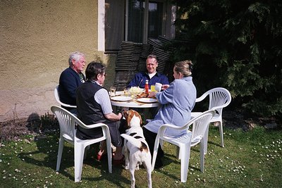 Four adults and a dog share a casual outdoor meal at a plastic table/chairs setup. Lighting suggests daytime, likely mid-20th...