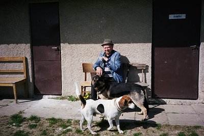 A man in a fedora and denim jacket sits on a wooden chair beside a rustic building, flanked by two dogs—one black-and-tan and...
