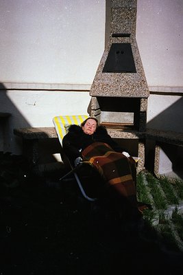 A woman reclines on a striped wooden bench beside a stone fireplace in an indoor courtyard. She wears a dark coat with a fur ...