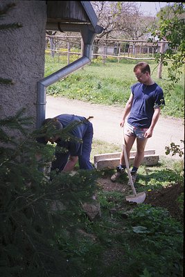 Two men gardening in a rural setting, 1970s. One kneels with shovel, the other stands holding a rake. Grey concrete house wit...