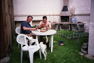 Two men seated at a white plastic table in a courtyard, sharing a meal with a dog nearby. The man on the left wears a dark sh...