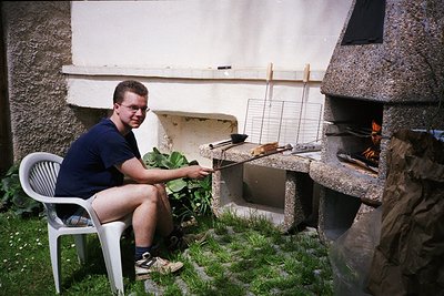 Man grills skewered meat over an open fire in a backyard setting, seated on a plastic chair. Mid-1990s outdoor cooking style ...
