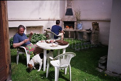 Two men relax in a courtyard with a brick oven, sharing a meal outdoors. One sits at a small round table with plates, cups, a...