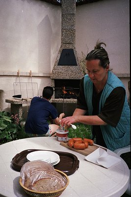 Indoor kitchen scene featuring two people preparing food by a stone fireplace. Woman in teal apron carves roasted meat on a c...