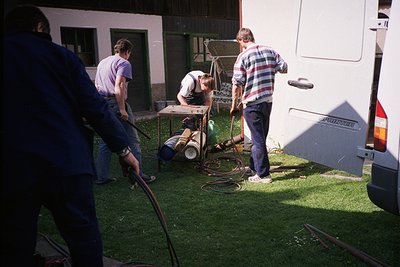 Three men in 1970s-style casual attire (plaid shirts, rolled-up jeans) operate a pressure washer on a white camper van, likel...