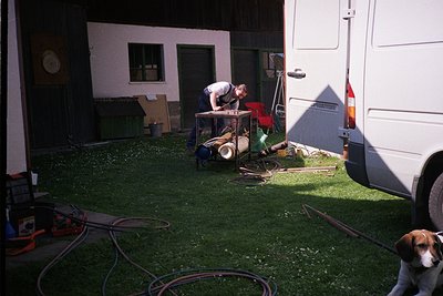 Two individuals work outdoors near a white RV parked beside a residential building. One person stands on a makeshift table, h...