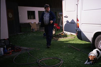 Mid-20th-century outdoor scene: man in plaid shirt and overalls walks beside a white RV, holding a hose. Lawn sprinkler coils...