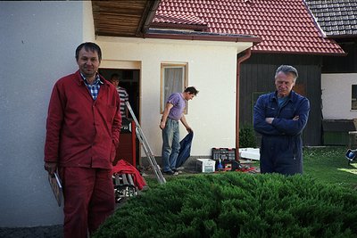Mid-20th century European home exterior with three men engaged in renovation. Left: man in red jacket and overalls holding a ...