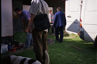 Group of three adults and a dog in a residential outdoor setting, likely a backyard. Two men in casual attire (one wearing a ...