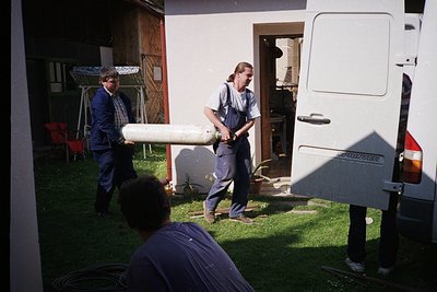 Two individuals transport a large cylindrical object (likely a telescope or antenna) near a white camper van (, , ). One pers...