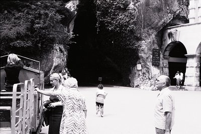 Mid-20th century black-and-white shot of a rocky coastal cave entrance with arched doorway. Tourists explore via metal railin...