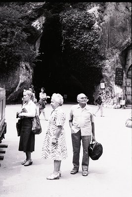 Three adults in mid-20th-century attire explore a cavern entrance, likely a tourist site. The man wears a patterned shirt and...