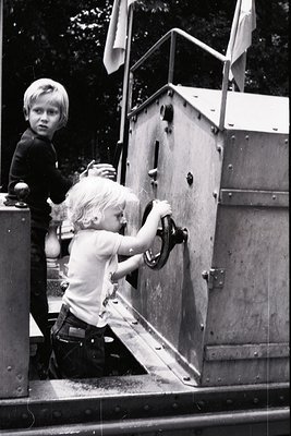Mid-20th century black-and-white photo of two children operating a vintage boat’s metal hatch. Boy in dark sweater leans over...