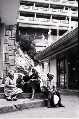 Mid-century urban courtyard with Brutalist concrete architecture. Five individuals—two adults, two children, and an elderly w...