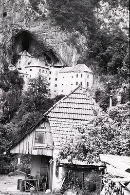 Historic alpine fortress perched on rocky cliffside, flanked by dense forest. Prominent medieval-style castle with arched win...