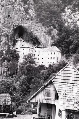Historic alpine fortress perched on a rocky cliffside, surrounded by dense forest. The multi-story stone structure features a...