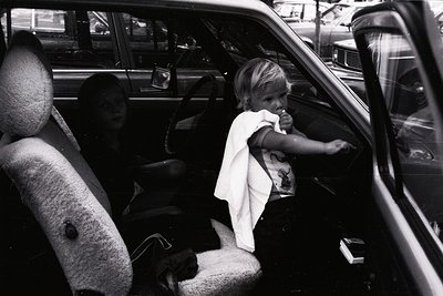 Vintage black-and-white street scene: young child in white dress leans out of classic car door, holding a snack. Adult seated...