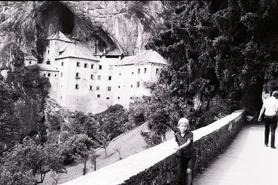A child stands on a narrow, stone-paved path beside a cliffside castle ruin, framed by dense forest. The castle’s multi-story...